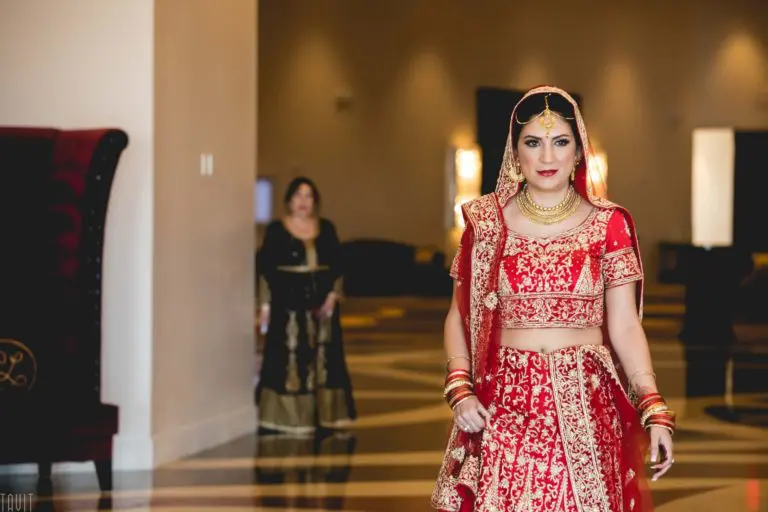 A woman dressed in a traditional red and gold bridal lehenga walks indoors, adorned with jewelry and a veil as if arriving at a seminar or corporate event. Another woman in a black and gold outfit stands in the background.