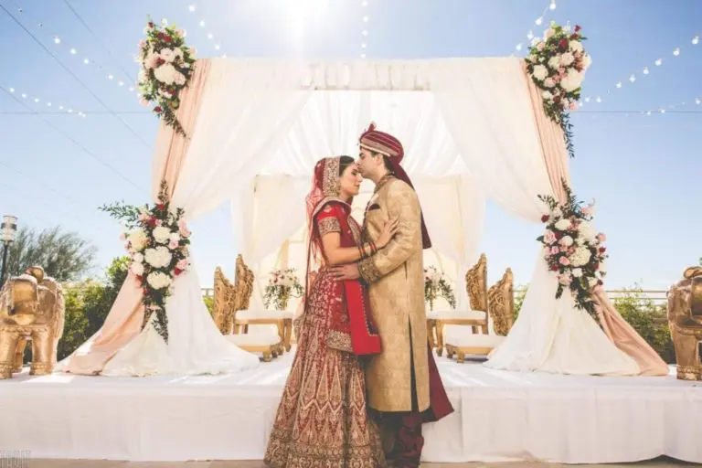 A bride and groom in traditional Indian wedding attire embrace on an outdoor decorated stage with floral arrangements, white drapery, and golden chairs—transforming the space that often hosts a seminar or corporate event into a magical celebration under a bright, sunny sky.