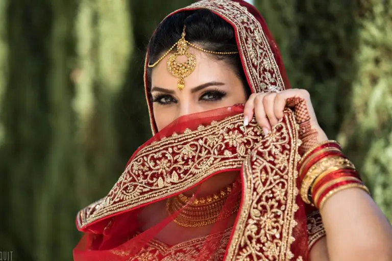 A woman in a red and gold traditional outfit covers her face with an ornate dupatta. She wears gold jewelry, including a maang tikka and bangles, standing gracefully as if attending a seminar, with greenery in the blurred background.