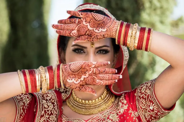 A bride in a red and gold traditional outfit poses with her henna-decorated hands framing her face, as if ready for a grand seminar or corporate event. She wears red bangles, gold jewelry, and a headpiece, with greenery blurred in the background.