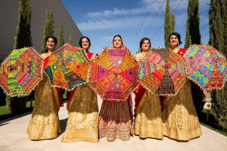 Five women in gold and red traditional outfits hold colorful, intricately patterned umbrellas, standing outside during a corporate event on a sunny day, with tall trees and a blue sky in the background.