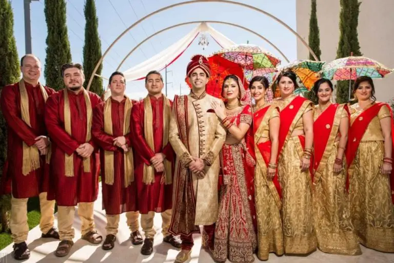 A wedding party poses outdoors as if at a festive corporate event; the groom and groomsmen wear red and gold attire, while the bride and bridesmaids don gold dresses with red accents. Two women hold colorful umbrellas amid green trees and a white arch.