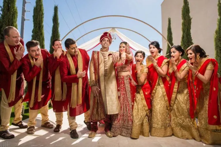 A bride and groom in traditional Indian wedding attire stand smiling, surrounded by joyful bridesmaids and groomsmen in gold and red outfits, all reacting enthusiastically outdoors under a decorated canopy at a festive seminar.