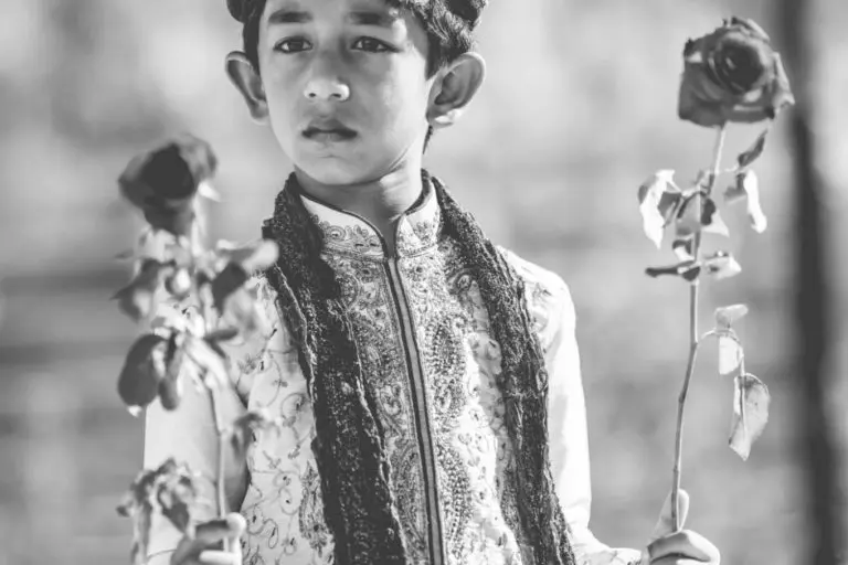 A young boy dressed in traditional attire and a decorative turban holds a rose in each hand, looking thoughtfully ahead—as if ready to welcome guests at a corporate event. The image is in black and white with a soft, blurred background.