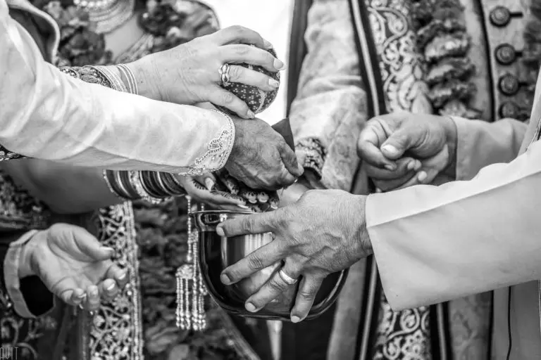 A close-up, black-and-white image shows several hands in ornate traditional clothing participating in a ceremonial ritual, pouring water from a vessel into a metal bowl at a corporate event.