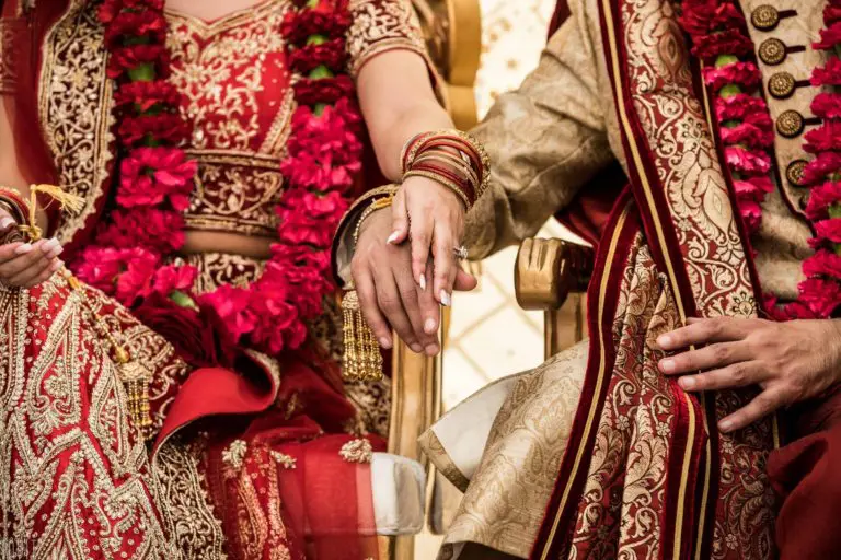 A close-up of a bride and groom in traditional Indian wedding attire, sitting side by side and holding hands, as if posing for a corporate event or seminar spotlight, adorned in red and gold outfits with intricate embroidery, jewelry, and floral garlands.
