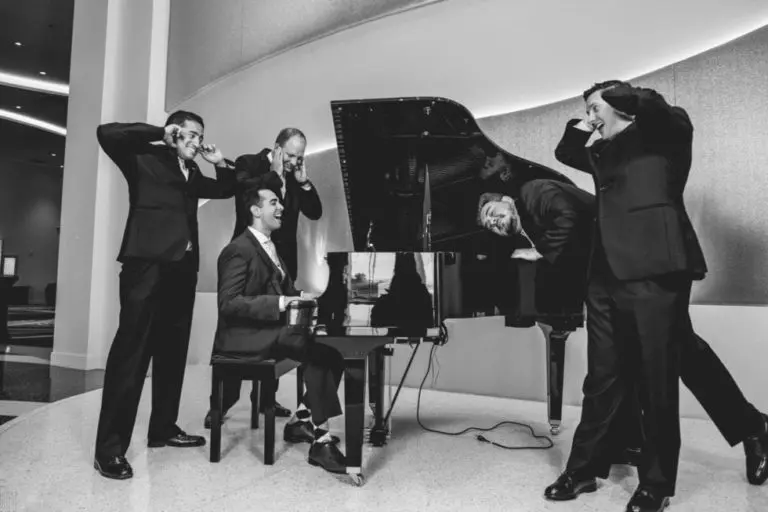 Four men in suits make funny faces and cover their ears at a corporate event while a fifth man, also suited, plays a grand piano. The scene unfolds indoors, captured in striking black and white.