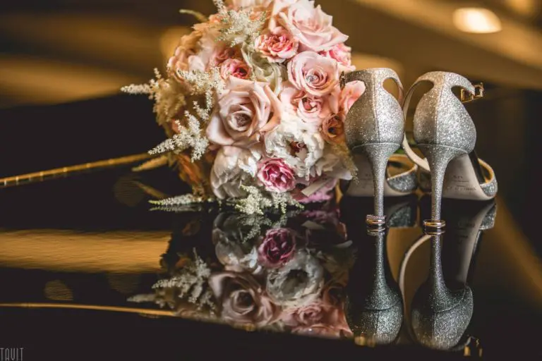 A bridal bouquet of pink and cream roses rests beside a pair of sparkling silver high-heeled shoes, both reflected on a glossy black surface—perfect elegance for a wedding or stylish corporate event.
