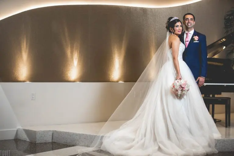 A bride in a white gown and veil holds a bouquet beside a groom in a blue suit with a pink boutonniere. They smile in a modern indoor setting, where soft, curved lighting evokes the elegance of a high-end seminar.