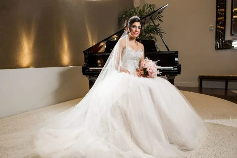 A bride in a white wedding gown and veil sits in front of a grand piano, holding a bouquet of light pink flowers and smiling, with an elegant interior that could also host a stylish corporate event in the background.