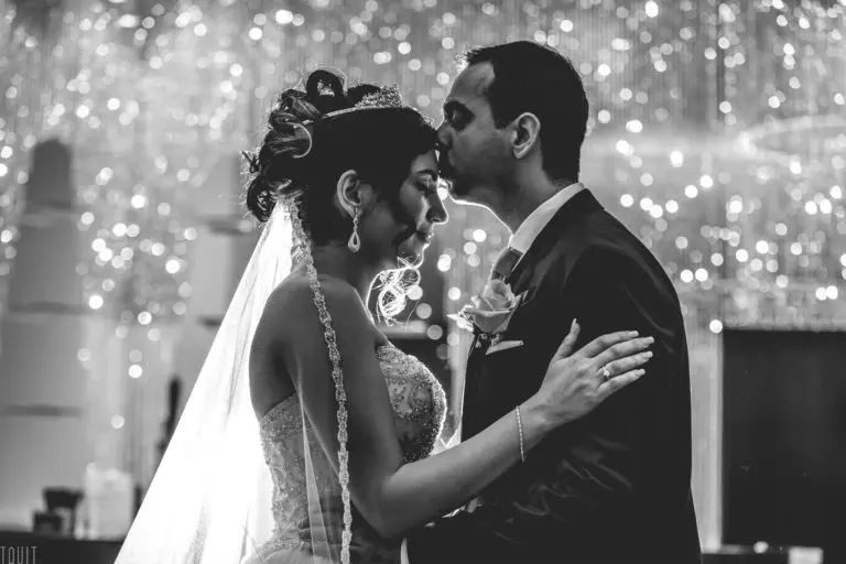 A groom gently kisses the forehead of his bride as they embrace on the dance floor, surrounded by sparkling lights in a romantic, elegant setting reminiscent of a beautifully decorated corporate event.
