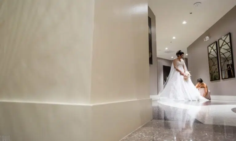 A bride in a white gown stands in a hallway, holding her bouquet, while another woman in a peach dress sits on the floor nearby. The foreground shows a large cream-colored column, evoking the elegance of a grand seminar or corporate event venue.