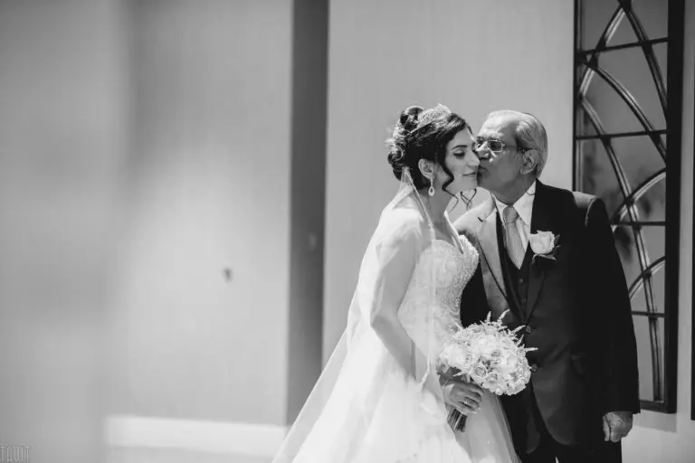 A bride in a wedding gown holds a bouquet while an older man in a suit kisses her cheek. They stand indoors at what could be a seminar or celebration; the scene is tender and joyful, captured in black and white.