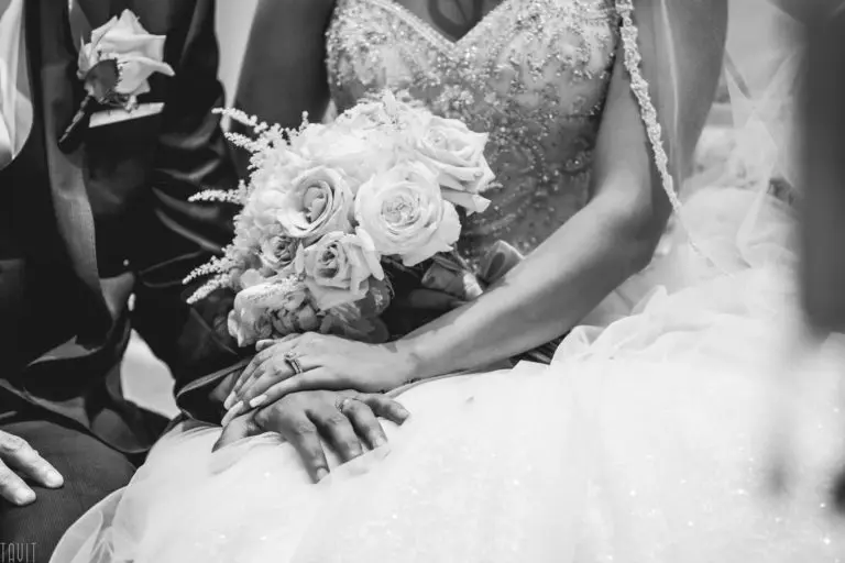 A black and white photo of a bride holding a bouquet of roses, resting her wedding-ringed hand atop the groom’s during a formal event, both elegantly dressed—capturing intimacy amid the atmosphere of a corporate event.