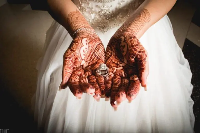 A bride in a white dress holds out her hands decorated with intricate mehndi designs, displaying a diamond ring in her palms, reminiscent of the grace seen at a formal seminar or corporate event.