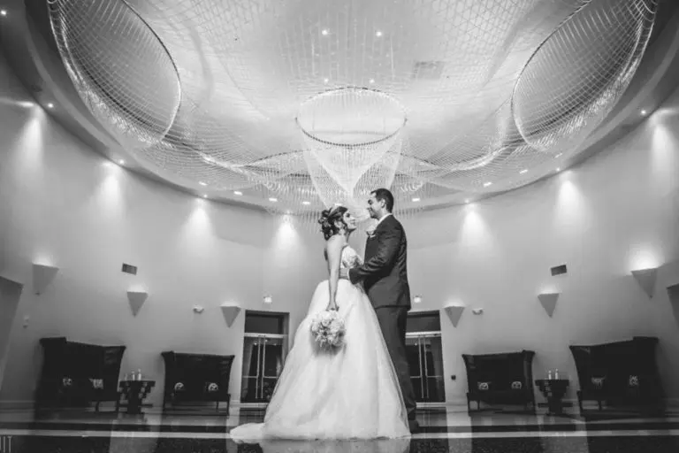 A bride and groom stand facing each other in an elegant, modern hall—often used for corporate events or seminars—holding hands and gazing into each other's eyes beneath a large decorative ceiling feature. The image is in black and white.