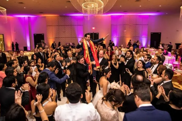 A joyous wedding reception with a large crowd dancing and celebrating, reminiscent of a lively corporate event. A man wearing a red and gold scarf is lifted on a chair above the crowd, under elegant chandeliers and colorful purple and orange lighting.