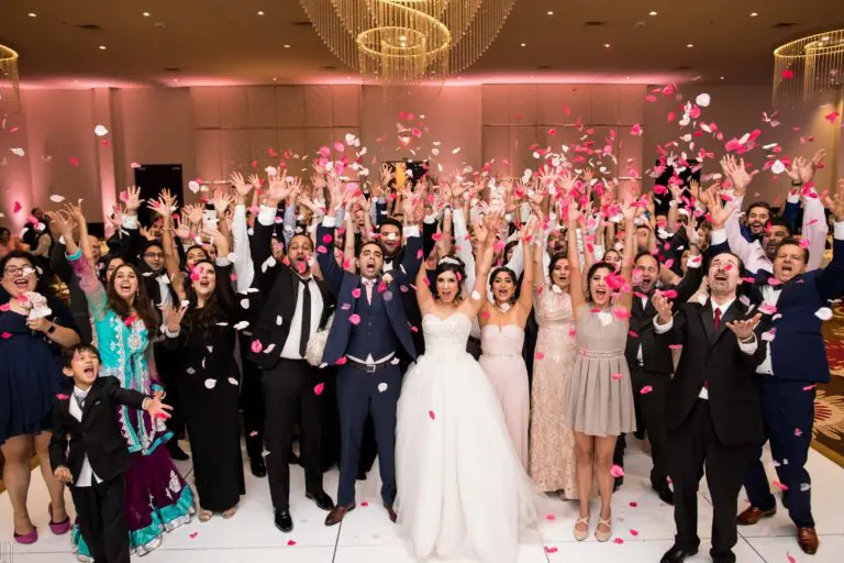 A large group of people dressed in formal attire, including a bride and groom at the center, smile and throw pink rose petals in the air during a lively indoor wedding celebration that has the elegance of a high-end corporate event.
