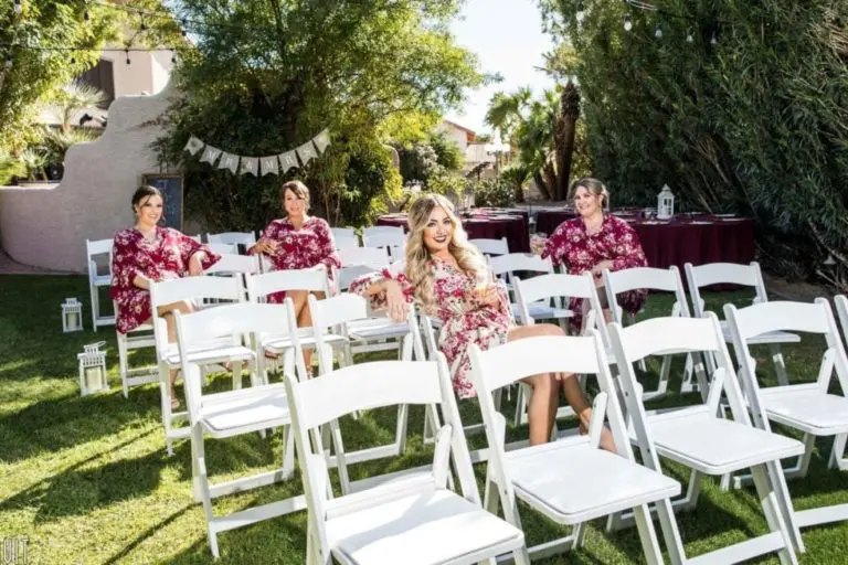 Four women in matching floral robes sit smiling on white folding chairs arranged outdoors, surrounded by greenery, likely preparing for a seminar or special event on a sunny day.