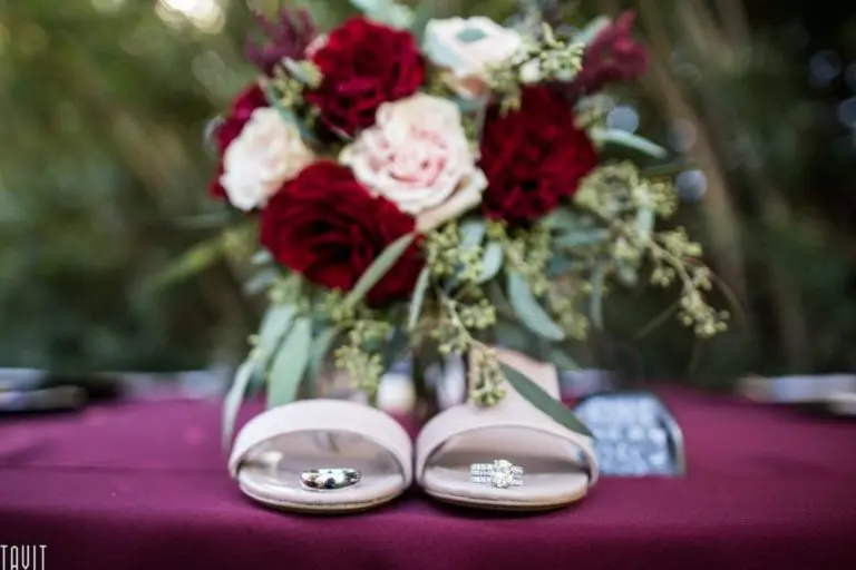 A pair of blush pink open-toe heels displayed in front of a bouquet of red and white roses, with rings and earrings on the shoes, set on a burgundy tablecloth outdoors—perfect for adding elegance to any corporate event.