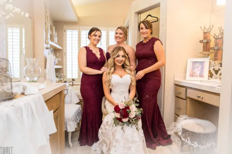 A bride in a white lace gown holds a bouquet while seated in a bright room, surrounded by three bridesmaids in matching burgundy dresses, all smiling warmly at the camera as if posing for a joyful seminar or corporate event photo.