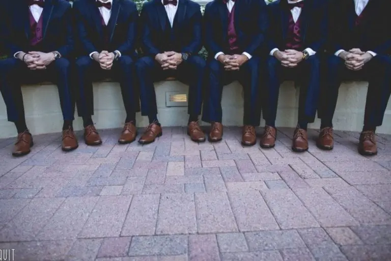 Six men dressed in dark suits, burgundy vests, and brown shoes sit side by side on a bench at a corporate event, hands clasped on their laps, with only their bodies visible from the neck down.