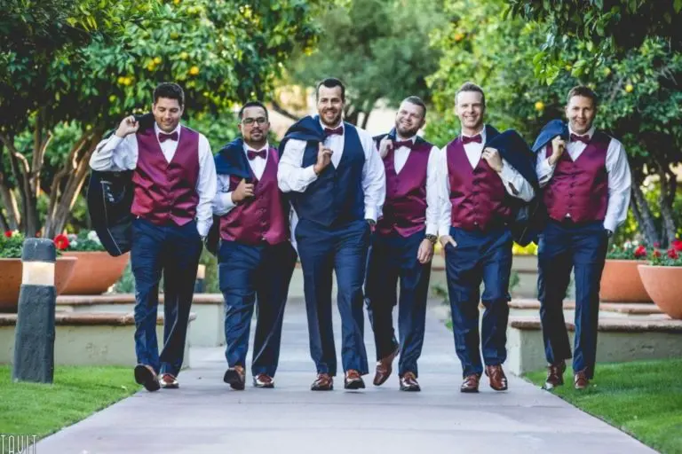 Six men dressed in matching navy suits and burgundy vests walk confidently outdoors on a pathway, as if heading to a seminar or corporate event. Trees and large planters with flowers line the path.