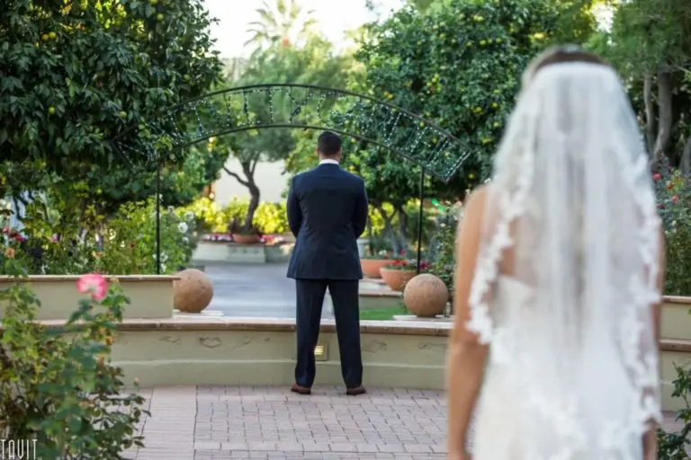 A bride in a white dress and veil approaches a groom in a suit, who stands with his back to her in a garden setting for their first look before the wedding, creating a romantic scene that contrasts beautifully with any formal seminar or corporate event.