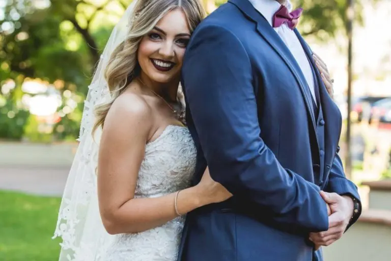 A bride in a white lace dress and veil stands closely behind a groom in a navy blue suit and red bow tie. Their warm smiles shine outdoors, with greenery and sunlight—turning their wedding into a moment more joyful than any corporate event or seminar.