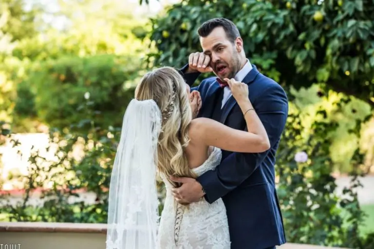 A bride and groom face each other outdoors, playfully pretending to box while smiling and laughing—adding a fun twist that would lighten up any seminar or corporate event. The bride wears a white gown; the groom is in a navy suit. Green trees blur behind them.