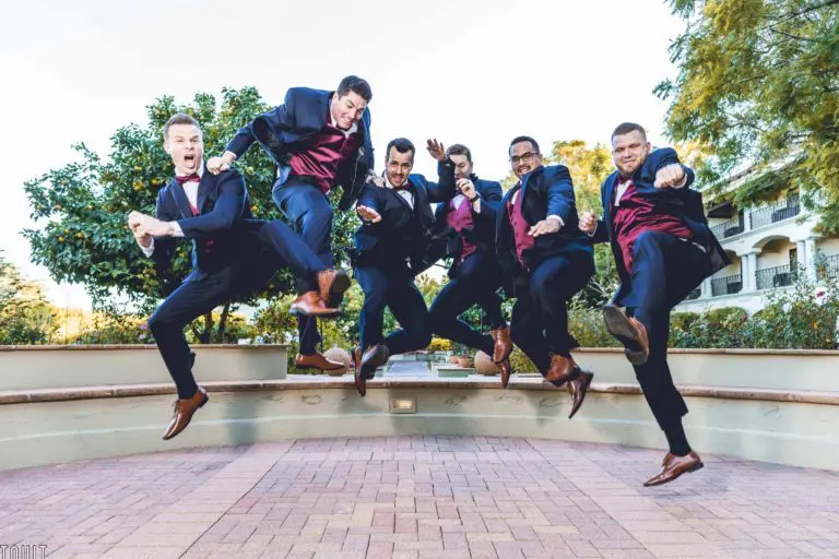 Six men in suits and burgundy vests jump simultaneously in mid-air at a corporate event, striking playful and energetic poses outdoors, with trees and a building in the background.