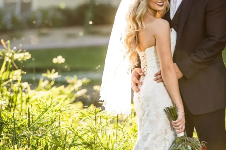 A bride in a lace gown and veil holds a bouquet while smiling at the camera, as the groom in a dark suit gently embraces her outdoors in golden sunlight—capturing romance far from any seminar or corporate event, surrounded by lush greenery.