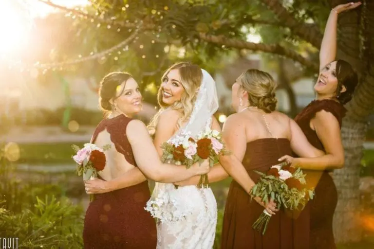 A bride in a white lace gown and veil poses outdoors with three bridesmaids in burgundy dresses—radiant as if attending a corporate event—smiling and holding bouquets, with sunlight streaming through trees in the background.