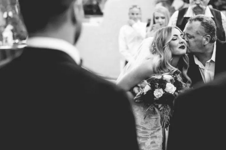 A bride holding a bouquet leans in to receive a kiss on the cheek from an older man, likely her father, during an outdoor wedding ceremony. Two people are seen in the blurred foreground, resembling candid moments at a seminar. The photo is in black and white.