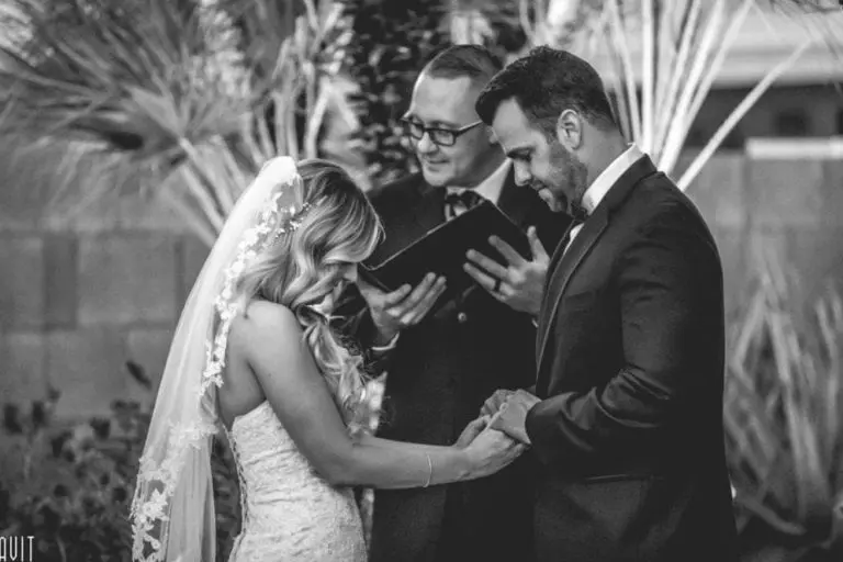 A bride and groom hold hands and look down during an outdoor wedding ceremony, as an officiant—reminiscent of a seminar or corporate event speaker—stands behind them reading from a book. The scene is in black and white.