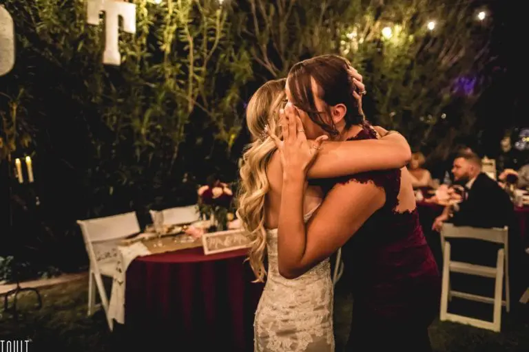 Two women in formal dresses embrace emotionally at an outdoor event, possibly a seminar or wedding reception, with decorated tables and string lights enhancing the atmosphere.