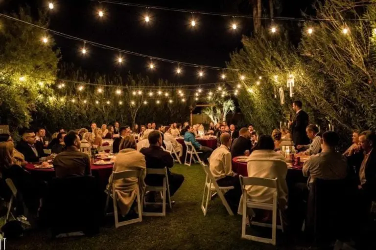 A large group of people sit at round tables with white chairs under string lights at an outdoor evening corporate event surrounded by trees, listening to a speaker standing at the front.