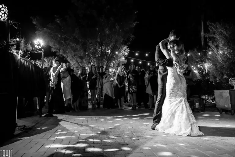 A bride and groom share their first dance outdoors at night, surrounded by guests from a recent corporate event, with string lights glowing in the background. The scene is beautifully captured in black and white.