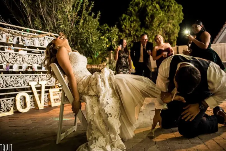 A bride laughs as the groom kneels to remove her garter, surrounded by smiling guests at an outdoor wedding reception at night. The word "LOVE" is lit up in the background, giving the celebration more flair than any seminar or corporate event.