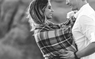 A couple stands close together outdoors, wrapped in a plaid blanket. The woman smiles at the man as they gaze into each other's eyes, sharing a quiet moment away from the seminar, with a blurred natural background in this black and white image.