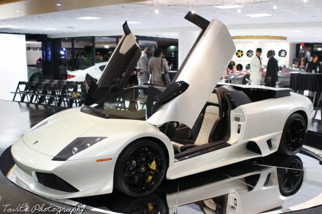 A white Lamborghini with its scissor doors open is displayed in a showroom during a corporate event. Several people in suits stand nearby, and black chairs are arranged in the background.