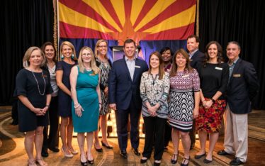 A group of thirteen people smile and pose together in front of a large Arizona state flag indoors, dressed in business or semi-formal attire. They appear to be attending a corporate event or ceremony.