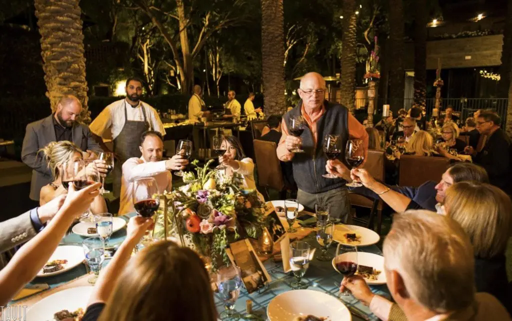 A group of people, captured in stunning corporate event photography, sits around a beautifully decorated dining table and raises their glasses for a toast. The table is set with plates and floral arrangements, while an outdoor setting with lush trees and soft lighting enhances the ambiance.