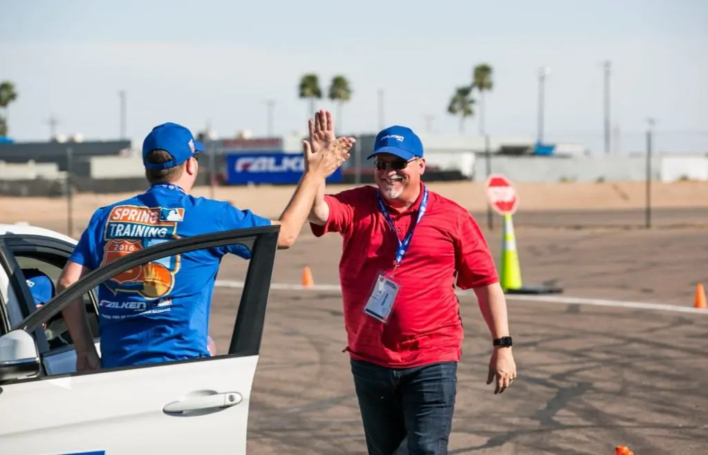 Two men, one in a red shirt and the other in blue, high-five next to a white car at a corporate event in a parking lot with traffic cones. Both are smiling, wearing caps and event lanyards.