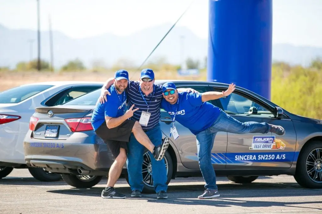 Three men in blue shirts and hats pose playfully in front of two parked cars with Falken branding at an outdoor corporate event, smiling and appearing cheerful under a sunny sky.