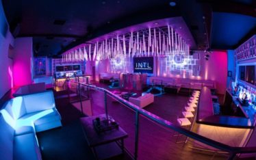 A modern nightclub interior with vibrant purple and blue lighting serves as an ideal venue for corporate event photography. The space features white sofas, a bar area with high stools, a dance floor, and decorative hanging light fixtures, creating a sleek and futuristic ambiance.