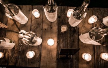 A rustic wooden table, ideal for corporate event photography, is adorned with small lit candles and glass bottles, viewed from above. The warm glow from the candles creates a cozy and inviting atmosphere.