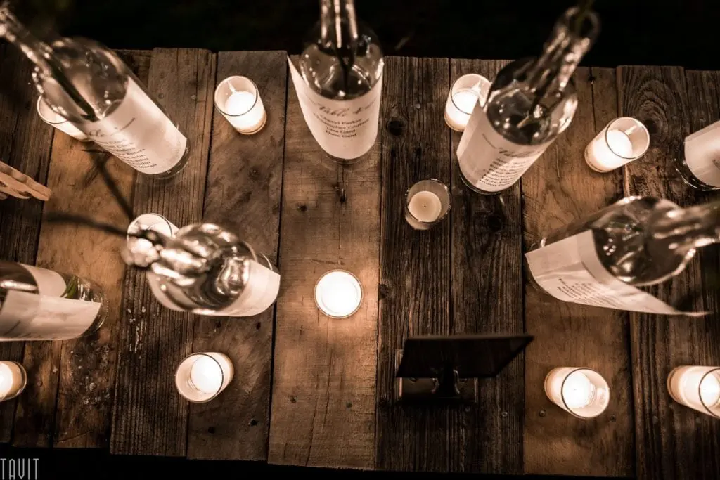 A rustic wooden table adorned with lit candles and translucent bottles, casting a warm, inviting glow perfect for corporate event photography. The bottles provide a reflective surface, enhancing the cozy ambiance ideal for capturing authentic moments.