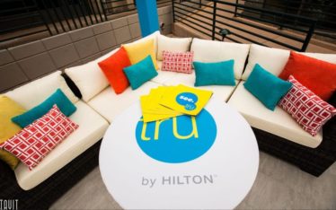 A curved outdoor sofa with white cushions and colorful pillows surrounds a round table featuring the “tru by Hilton” logo and several bright yellow folders, perfect for a relaxed corporate event or seminar setting.