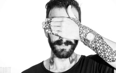 A man with a beard covers his eyes with his tattooed hand, featuring geometric patterns. He wears a watch, rings, and a chain necklace—an intriguing black-and-white portrait suited for a seminar or corporate event backdrop.